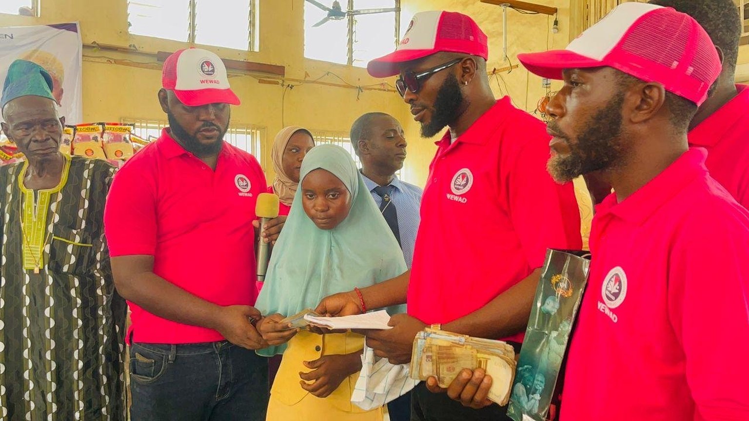 WEWAD volunteers in branded shirts and caps hand support to a student at a community distribution in Nigeria, with bagged provisions stacked behind them.